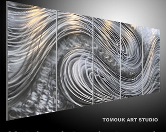 May include: A silver metal sculpture with a swirling, abstract design. The sculpture is made up of multiple panels that are joined together. The sculpture is lit from the front, highlighting the details of the design.