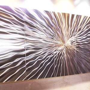 May include: A large, abstract metal wall sculpture with a silver finish. The sculpture is made up of multiple panels that are joined together to create a flowing, wave-like pattern.