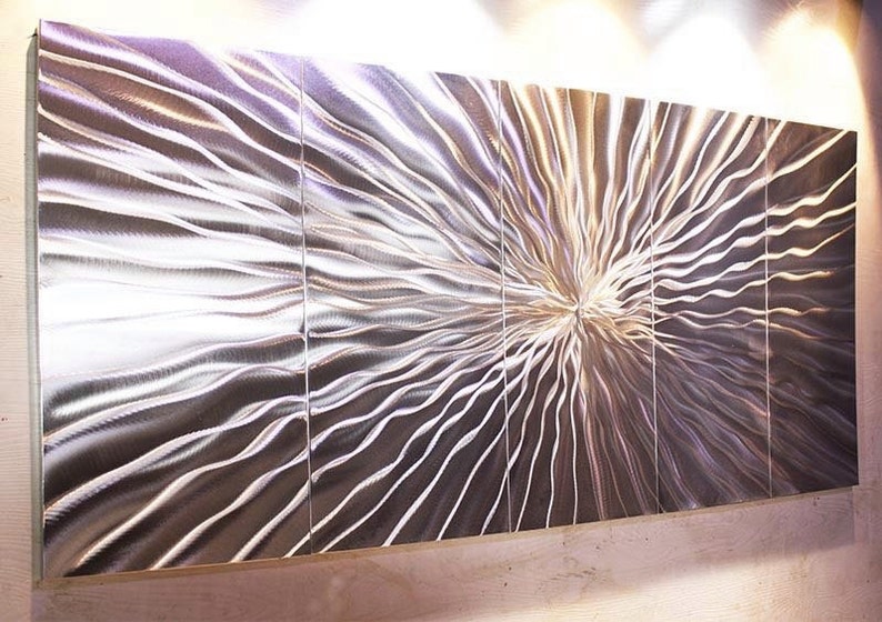 May include: A large, abstract metal wall sculpture with a silver finish. The sculpture is made up of multiple panels that are joined together to create a flowing, wave-like pattern.