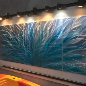 May include: A three-panel metal wall art piece with a blue and silver abstract design. The art is illuminated by track lighting.