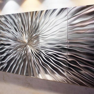 May include: A large, abstract metal wall sculpture with a silver finish. The sculpture is made up of multiple panels that are joined together to create a flowing, wave-like pattern.