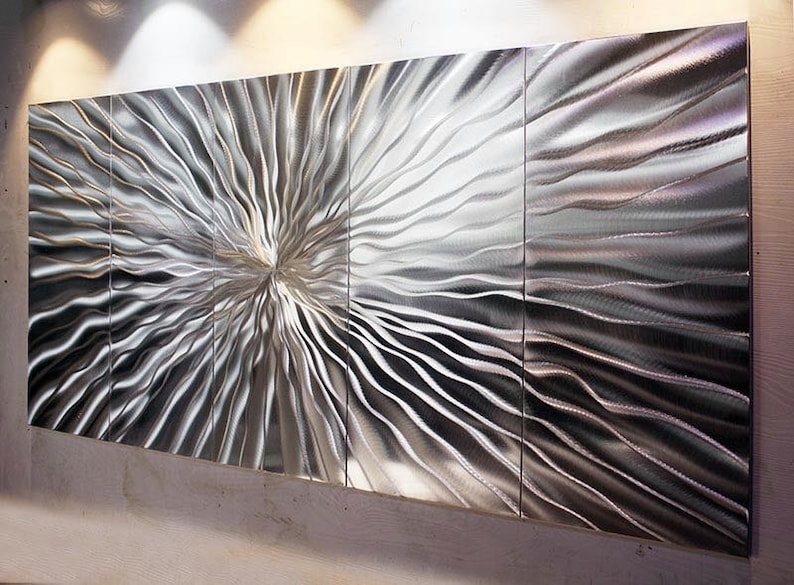 May include: A large, abstract metal wall sculpture with a silver finish. The sculpture is made up of multiple panels that are joined together to create a flowing, wave-like pattern.