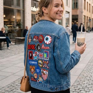 Könnte beinhalten: Eine blaue Jeansjacke mit einer Vielzahl bunter gestickter Aufnäher auf der Rückseite, darunter Logos von AC/DC, Yamaha, Vespa sowie verschiedene grafische Motive wie ein Comic-Auge, ein Tiger und ein Totenkopf, getragen mit einer beigen Umhängetasche.