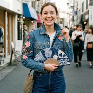 Puede incluir: Una chaqueta de mezclilla azul con parches bordados de rosas rosadas en la manga derecha y un parche en forma de corazón en el pecho izquierdo. La chaqueta se usa sobre una camisa blanca, combinada con jeans azules y un bolso cruzado beige.