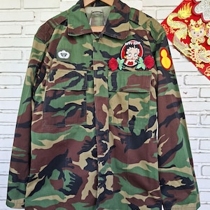May include: A camouflage print long-sleeve jacket featuring a Betty Boop patch with two red roses on the left chest pocket, a small white parachute emblem on the right chest, and a yellow oval patch on the left sleeve, all on a green, brown, and black pattern.