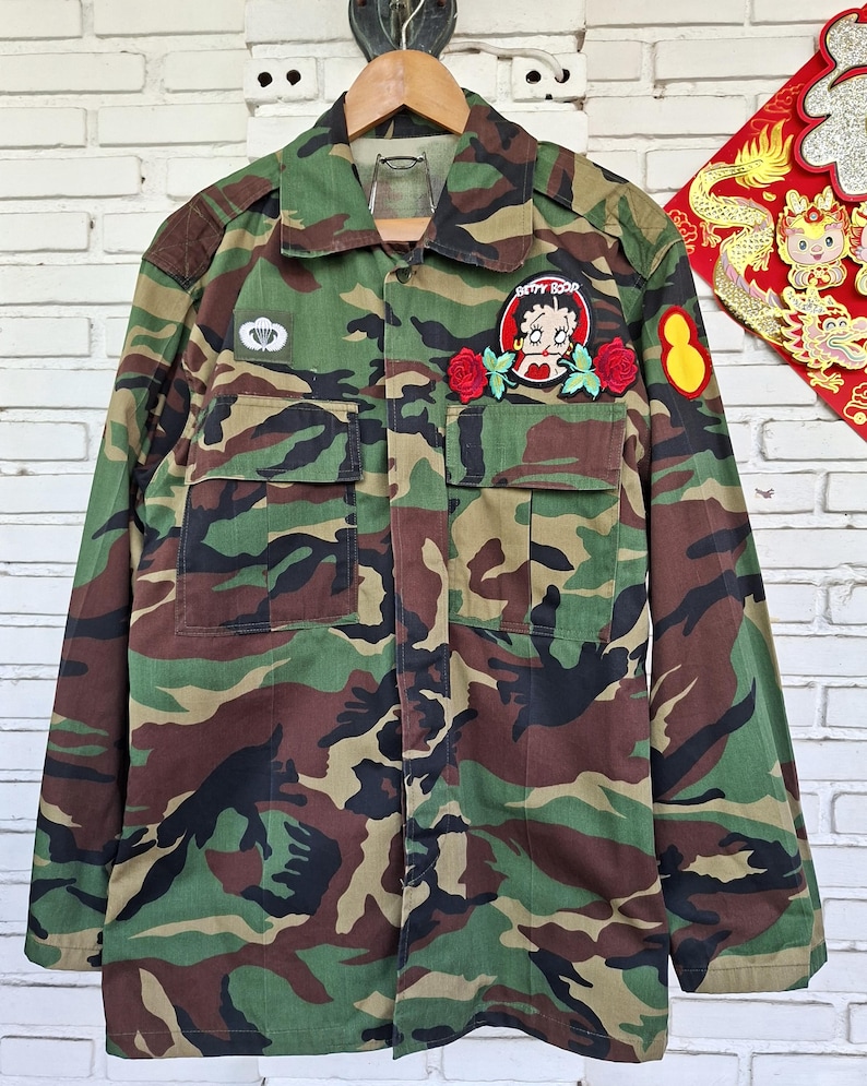 May include: A camouflage print long-sleeve jacket featuring a Betty Boop patch with two red roses on the left chest pocket, a small white parachute emblem on the right chest, and a yellow oval patch on the left sleeve, all on a green, brown, and black pattern.