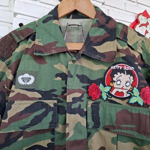 May include: A camouflage print long-sleeve shirt featuring a Betty Boop patch with red roses, a white parachute insignia patch on the right chest, and a yellow gourd-shaped patch on the left sleeve. The shirt has a collar and two front pockets.