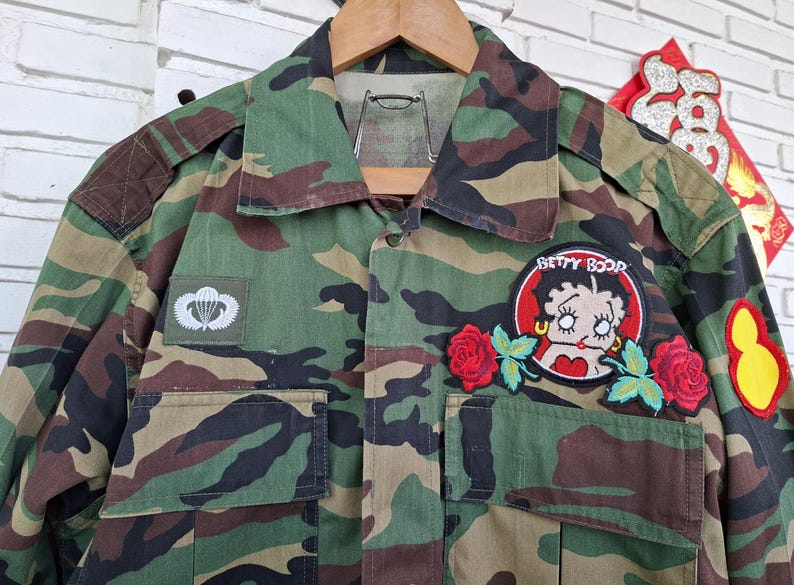 May include: A camouflage print long-sleeve shirt featuring a Betty Boop patch with red roses, a white parachute insignia patch on the right chest, and a yellow gourd-shaped patch on the left sleeve. The shirt has a collar and two front pockets.
