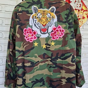 May include: A camouflage print jacket featuring a large embroidered tiger head patch on the back, flanked by two pink floral patches. Below the tiger, three small yellow embroidered stars are arranged in a horizontal row on the fabric.