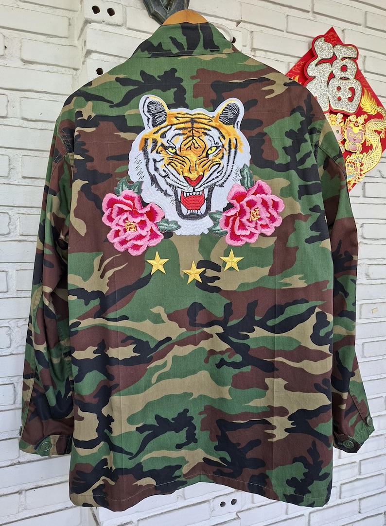 May include: A camouflage print jacket featuring a large embroidered tiger head patch on the back, flanked by two pink floral patches. Below the tiger, three small yellow embroidered stars are arranged in a horizontal row on the fabric.
