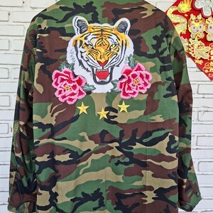 May include: A camouflage print jacket featuring a large embroidered tiger head patch on the back, flanked by two pink peony flower patches. Below the tiger, three small yellow embroidered stars are arranged in a horizontal row on the fabric.