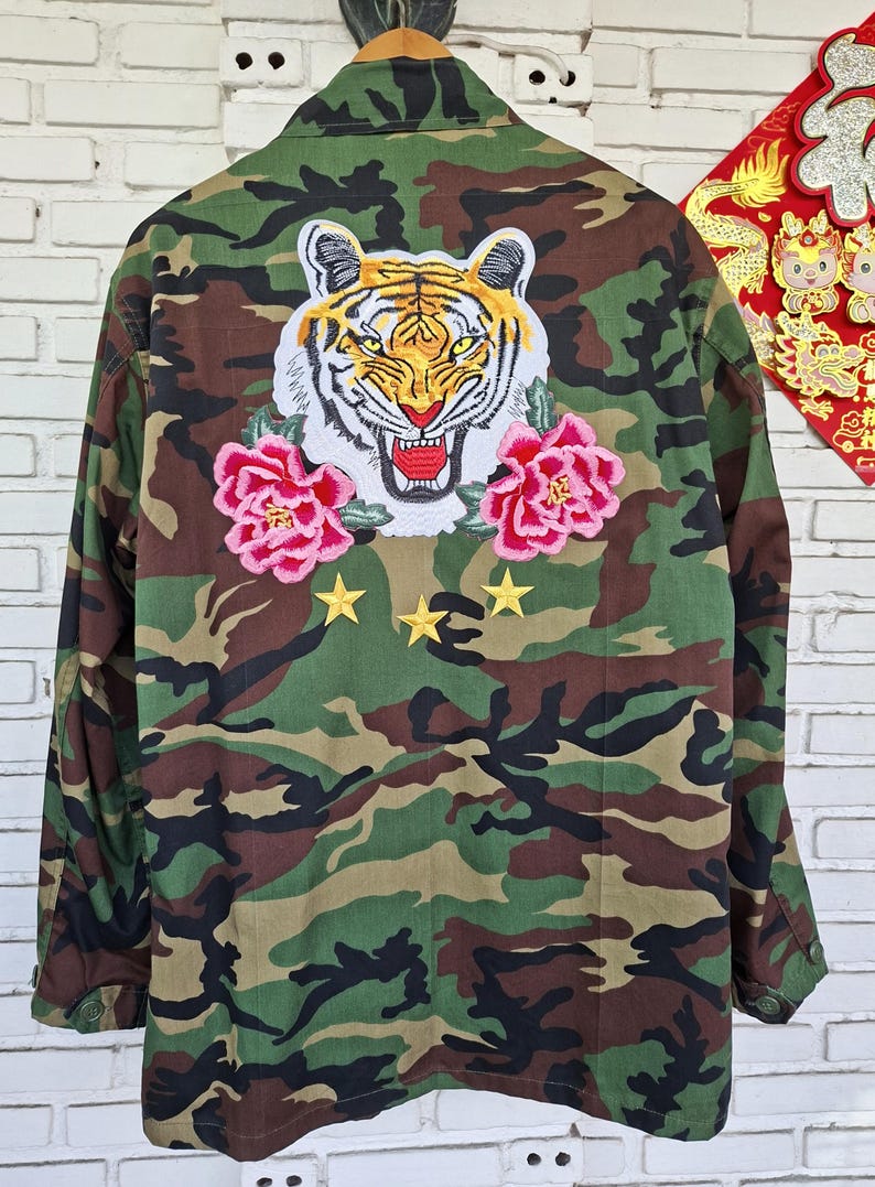 May include: A camouflage print jacket featuring a large embroidered tiger head patch on the back, flanked by two pink peony flower patches. Below the tiger, three small yellow embroidered stars are arranged in a horizontal row on the fabric.