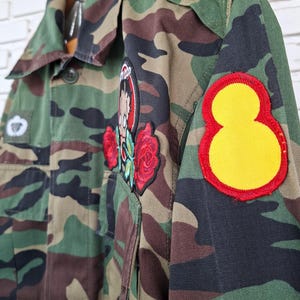 May include: A camouflage print jacket featuring a large circular patch on the sleeve with a yellow center and red border. The front includes a patch of a cartoon character surrounded by two red embroidered roses and a small rectangular patch.