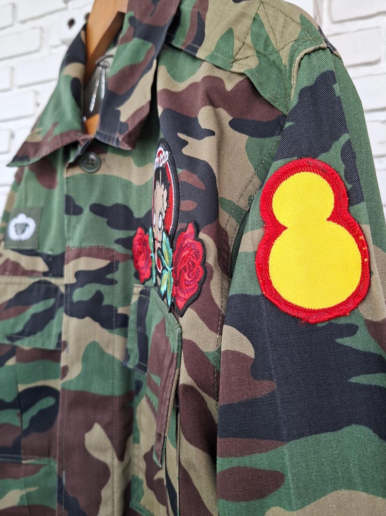 May include: A camouflage print jacket featuring a large circular patch on the sleeve with a yellow center and red border. The front includes a patch of a cartoon character surrounded by two red embroidered roses and a small rectangular patch.