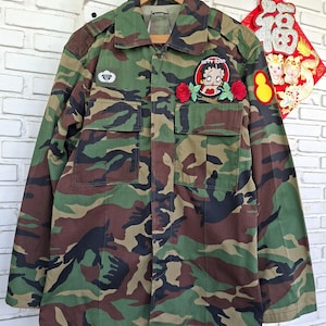 May include: A camouflage print long-sleeve button-up shirt featuring a collar, two chest pockets, and embroidered patches. The patches include a white parachute emblem, a Betty Boop character graphic flanked by two red roses, and a yellow oval.