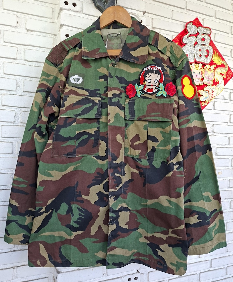 May include: A camouflage print long-sleeve button-up shirt featuring a collar, two chest pockets, and embroidered patches. The patches include a white parachute emblem, a Betty Boop character graphic flanked by two red roses, and a yellow oval.