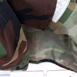 May include: A jacket featuring a woodland camouflage pattern in shades of dark brown, black, olive green, and tan. The garment has a visible white interior label with printed text, stitched seams, and a folded fabric construction showing the pattern.
