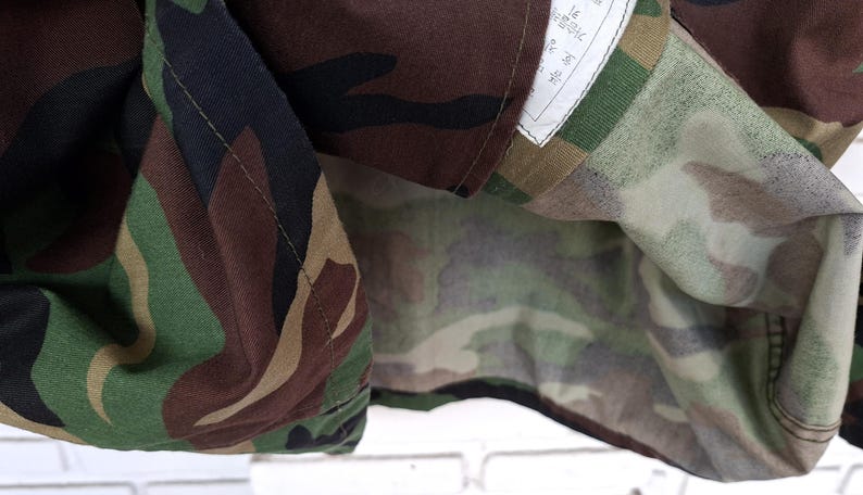 May include: A jacket featuring a woodland camouflage pattern in shades of dark brown, black, olive green, and tan. The garment has a visible white interior label with printed text, stitched seams, and a folded fabric construction showing the pattern.
