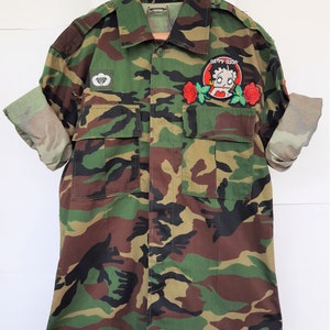 May include: A camouflage print button-down shirt with long sleeves rolled up. The shirt features a white parachute emblem on the right chest and a circular patch on the left chest depicting Betty Boop surrounded by two red roses.