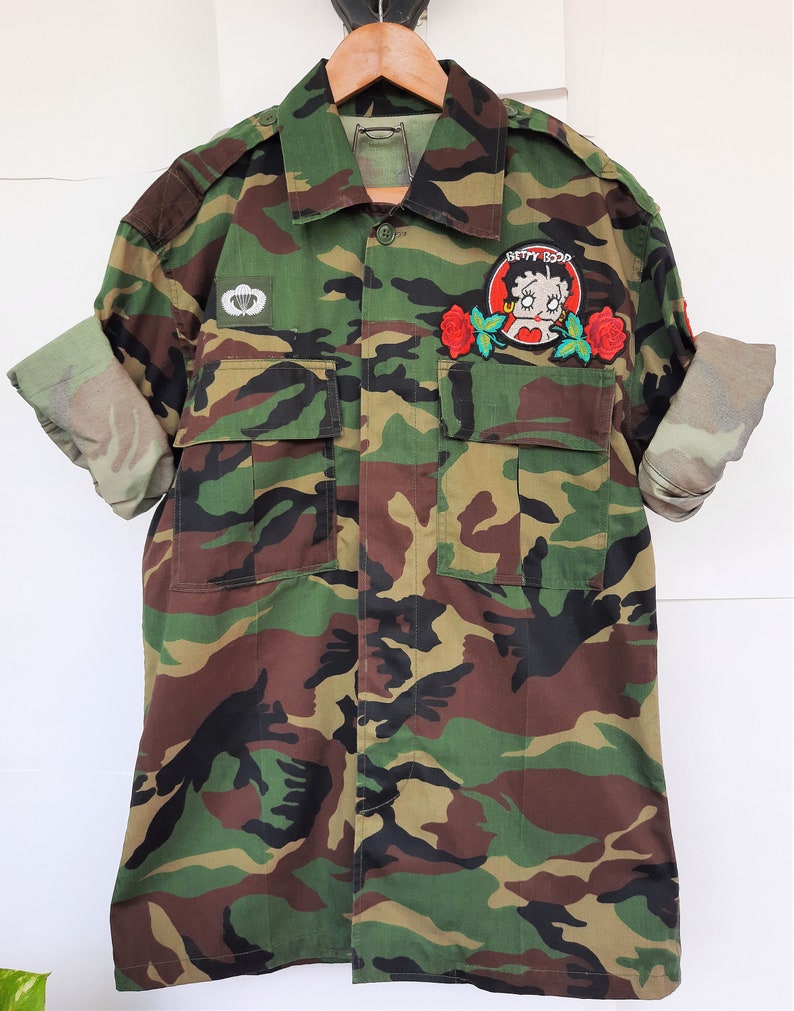 May include: A camouflage print button-down shirt with long sleeves rolled up. The shirt features a white parachute emblem on the right chest and a circular patch on the left chest depicting Betty Boop surrounded by two red roses.