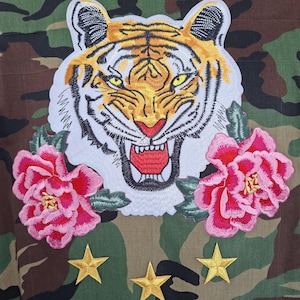 May include: A camouflage jacket featuring a large embroidered tiger head patch with yellow eyes and an open mouth, flanked by two pink peony flower patches. Below the tiger and flowers are three embroidered gold stars on the fabric.