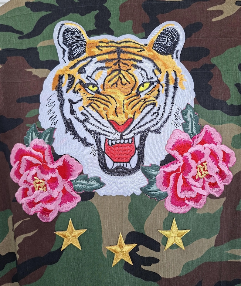 May include: A camouflage jacket featuring a large embroidered tiger head patch with yellow eyes and an open mouth, flanked by two pink peony flower patches. Below the tiger and flowers are three embroidered gold stars on the fabric.