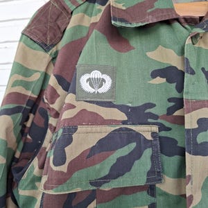 May include: A long-sleeve camouflage jacket featuring a green, brown, and black woodland pattern. The jacket includes a pointed collar, a button-down front, and a rectangular patch on the chest displaying a white embroidered parachute emblem.