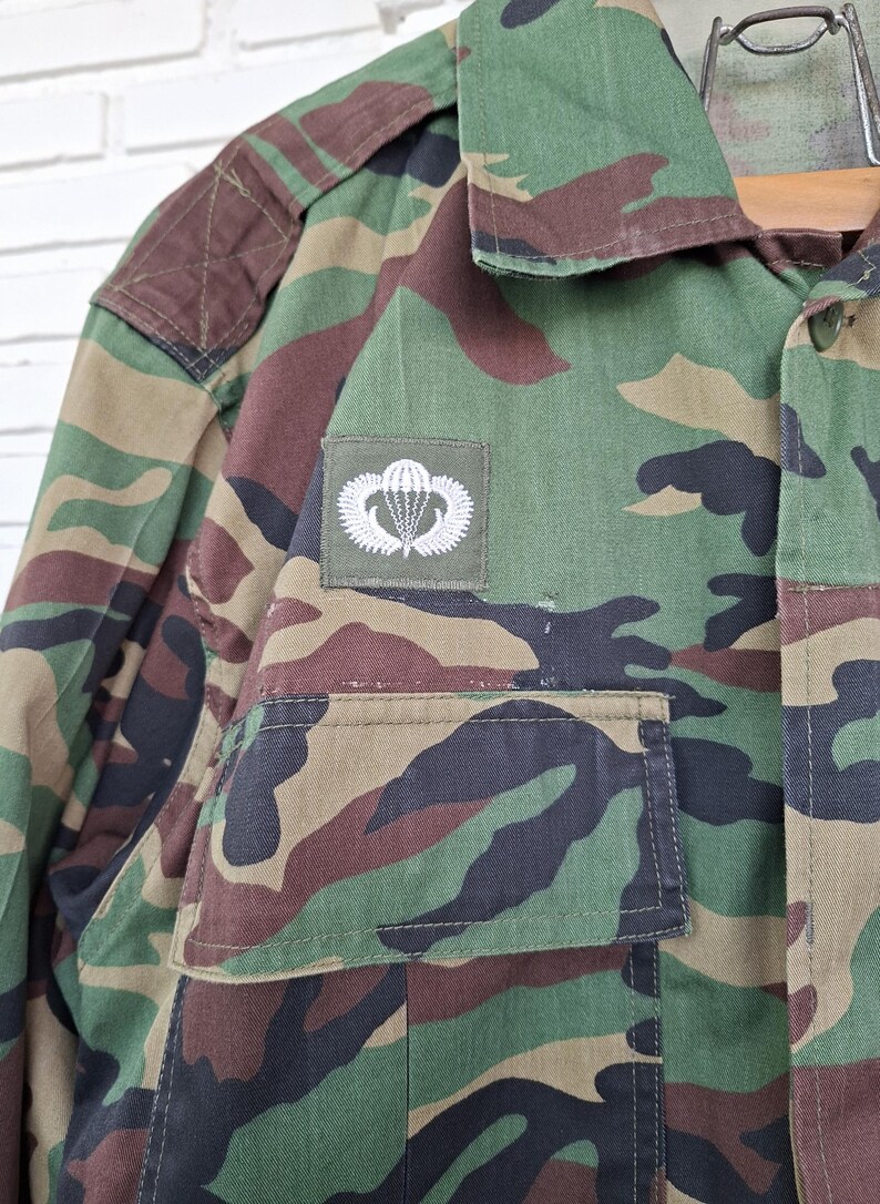 May include: A long-sleeve camouflage jacket featuring a green, brown, and black woodland pattern. The jacket includes a pointed collar, a button-down front, and a rectangular patch on the chest displaying a white embroidered parachute emblem.