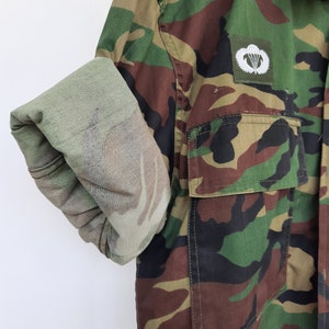 May include: A long-sleeve camouflage shirt featuring a woodland pattern of green, brown, and black. The shirt has a buttoned chest pocket and a white embroidered parachute emblem on the chest. One sleeve is rolled up to reveal the lighter interior fabric.