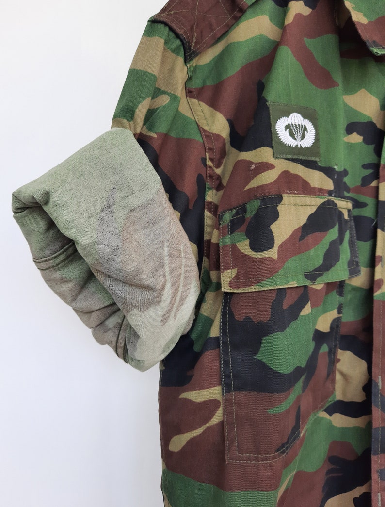 May include: A long-sleeve camouflage shirt featuring a woodland pattern of green, brown, and black. The shirt has a buttoned chest pocket and a white embroidered parachute emblem on the chest. One sleeve is rolled up to reveal the lighter interior fabric.