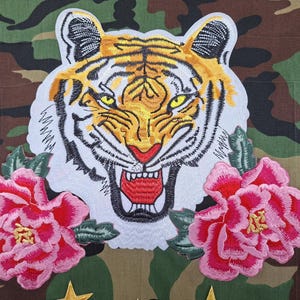 May include: A camouflage fabric garment featuring a large embroidered tiger head patch with yellow, black, and white fur, yellow eyes, and a red mouth. Two vibrant pink peony flower patches with green leaves are positioned below the tiger head.