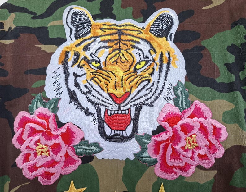 May include: A camouflage fabric garment featuring a large embroidered tiger head patch with yellow, black, and white fur, yellow eyes, and a red mouth. Two vibrant pink peony flower patches with green leaves are positioned below the tiger head.