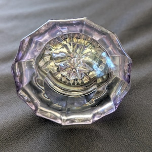 May include: A vintage, faceted glass doorknob in a soft lavender hue. The knob has a starburst design in the center and a multi-sided, octagonal shape. The glass reflects light, creating a shimmering effect.