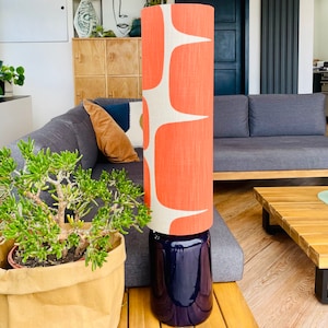 May include: A modern floor lamp with a coral orange and white patterned lampshade and a dark blue ceramic base. The lamp is sitting on a wooden table.