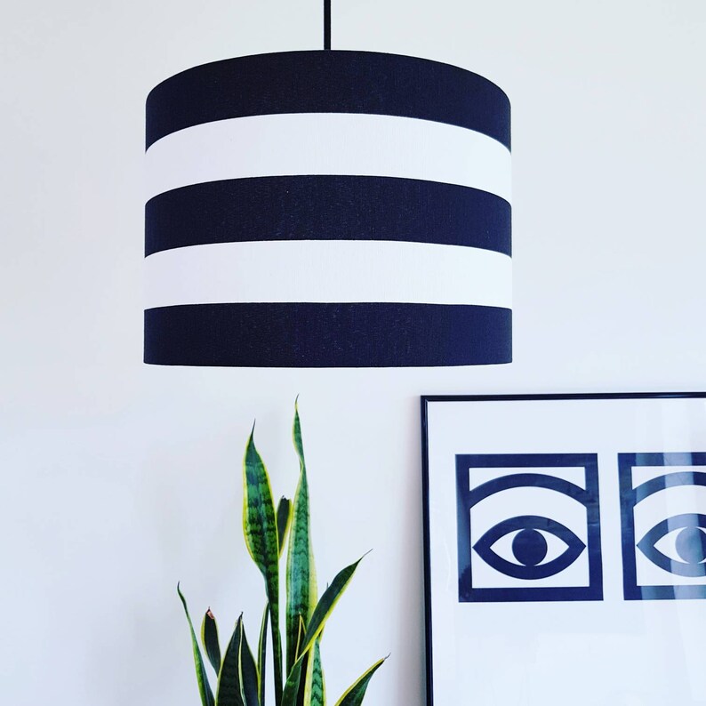 May include: A black and white striped lampshade with a black cord hanging from the ceiling. The lampshade is hanging over a green plant and a framed print with two black eyes.
