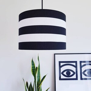 May include: A black and white striped lampshade hangs from a white ceiling. The lampshade is cylindrical and has three white stripes.