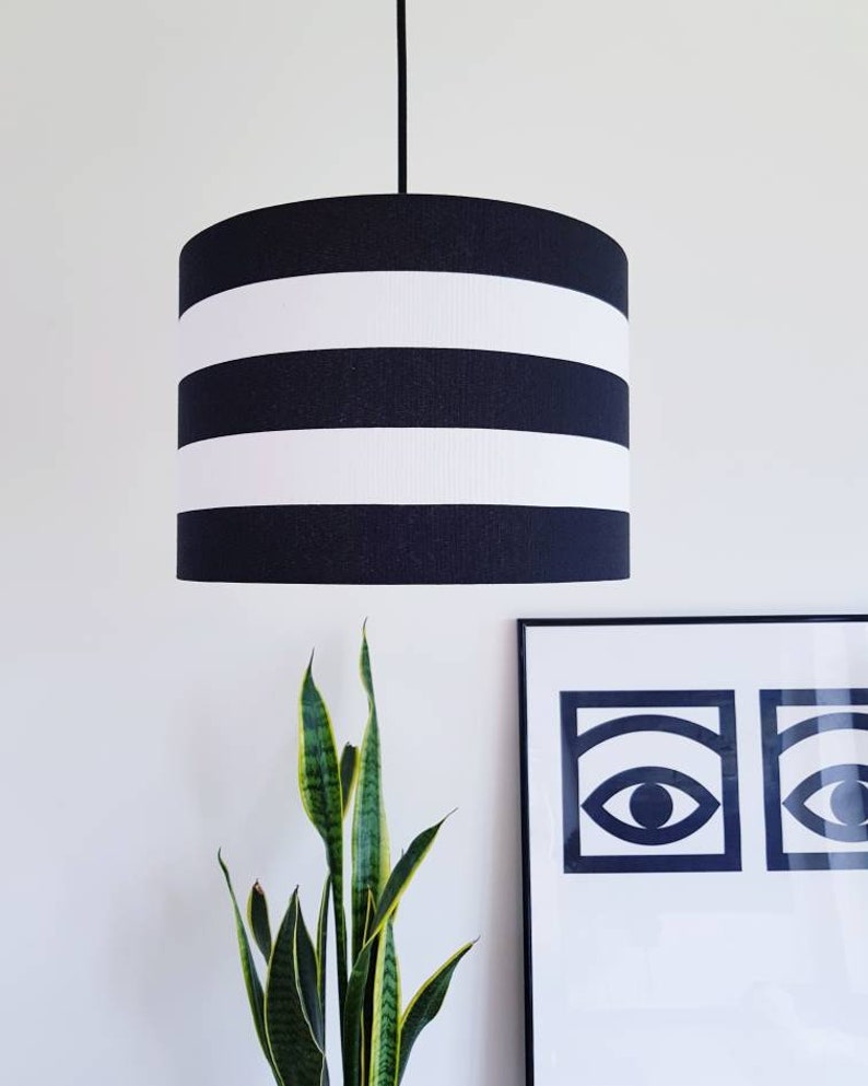 May include: A black and white striped lampshade hangs from a white ceiling. The lampshade is cylindrical and has three white stripes.