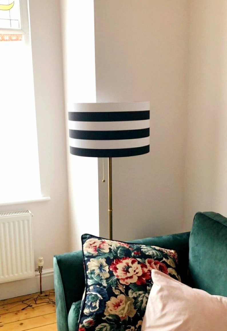 May include: A black and white striped lampshade with a gold metal base. The lamp is sitting on a green sofa with floral patterned pillows.