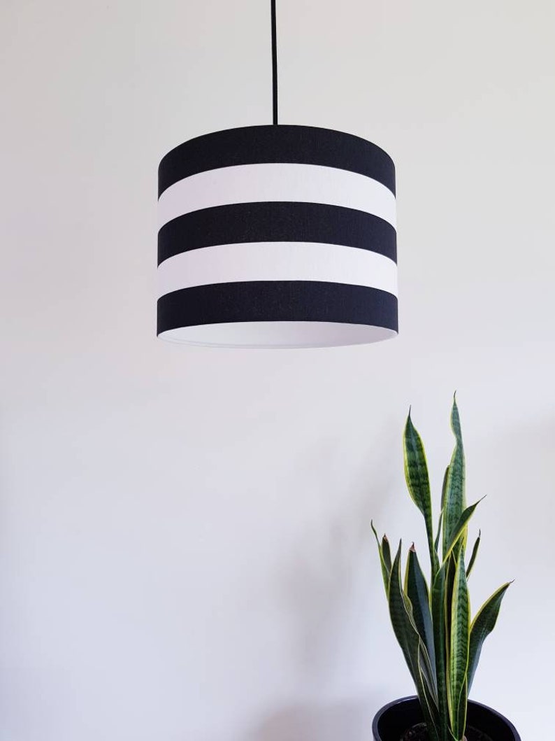 May include: A black and white striped lampshade hangs from a black cord against a white wall. A green plant is in the background.