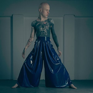 Pode incluir: Uma pessoa com a cabeça raspada e tatuagens está vestindo uma calça larga de cor azul escura brilhante com pregas. A calça é feita de um material liso e brilhante.