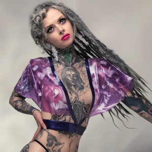 Könnte beinhalten: Eine Frau trägt ein transparentes, lila Oberteil mit Blumenmuster, dunkelblauen Besatz und einen schwarzen String. Sie trägt silberne Plateau-High Heels. Ihr Körper ist mit Tätowierungen bedeckt.