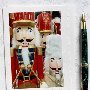 May include: A white greeting card featuring a photograph of several red and gold nutcracker figures, with the handwritten text Nutcrackers on Parade Kim Pletycha at the bottom, placed next to a green marbled fountain pen with gold accents.