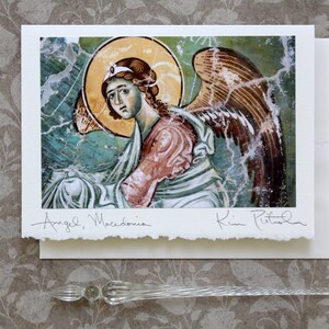 May include: A print of an angel from Macedonia, featuring a halo and wings, with the text "Angel, Macedonia" and a signature. The print is on textured paper, accompanied by an envelope and a glass pen.