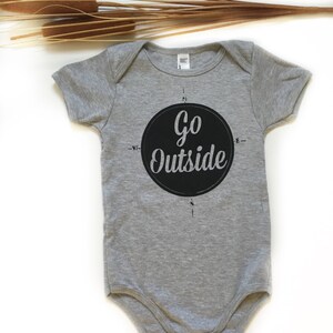 Pode incluir: Um macac&atilde;o de beb&ecirc; cinza com um design de b&uacute;ssola circular preto que diz "Go Outside" em texto branco.