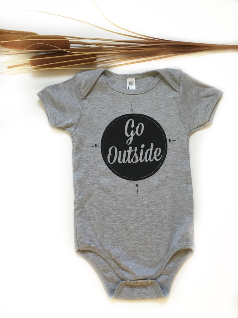 Pode incluir: Um macac&atilde;o de beb&ecirc; cinza com um design de b&uacute;ssola circular preto que diz "Go Outside" em texto branco.