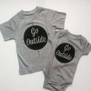 Pode incluir: Duas camisetas cinzas com estampa preta. A camiseta maior tem um design de b&uacute;ssola com as palavras "Go Outside" em um c&iacute;rculo. A camiseta menor tem o mesmo design, mas &eacute; um macac&atilde;o.