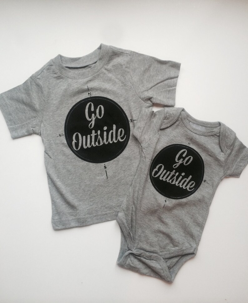 Pode incluir: Duas camisetas cinzas com estampa preta. A camiseta maior tem um design de b&uacute;ssola com as palavras "Go Outside" em um c&iacute;rculo. A camiseta menor tem o mesmo design, mas &eacute; um macac&atilde;o.