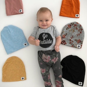 Pode incluir: Um beb&ecirc; vestindo um macac&atilde;o cinza com o texto "Go Outside" em um c&iacute;rculo preto, cal&ccedil;as cinzas florais e uma variedade de gorros de tric&ocirc; coloridos. Os gorros s&atilde;o nas cores azul claro, laranja, rosa, amarelo, cinza com estampa floral e preto.