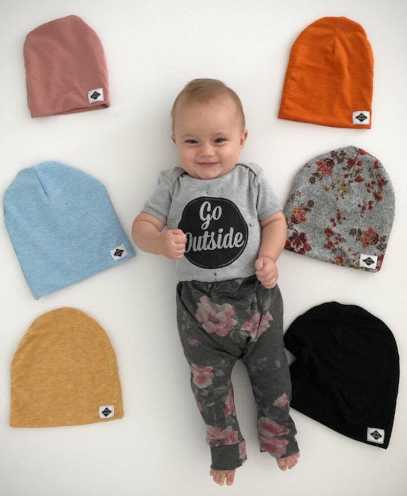 Pode incluir: Um beb&ecirc; vestindo um macac&atilde;o cinza com o texto "Go Outside" em um c&iacute;rculo preto, cal&ccedil;as cinzas florais e uma variedade de gorros de tric&ocirc; coloridos. Os gorros s&atilde;o nas cores azul claro, laranja, rosa, amarelo, cinza com estampa floral e preto.