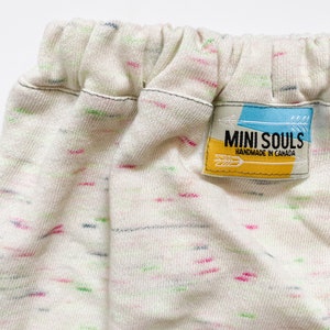 May include: White cotton baby trousers with a multi-coloured speckled pattern. The trousers have a black elastic waistband and a yellow label that reads "MINI SOULS HANDMADE IN CANADA".